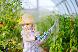 Adorable little girl wearing straw hat and childrens garden gloves playing with her toy garden tools in a greenhouse