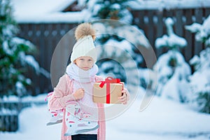 Adorable little girl wearing warm clothes outdoors on Christmas day holding gift and skates