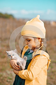 Adorable little girl in a yellow cap and raincoat holding kitty cat outdoor in nature. Pet concept