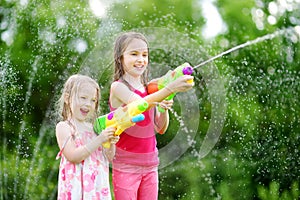 Adorable little girls playing with water guns on hot summer day. Cute children having fun with water outdoors.