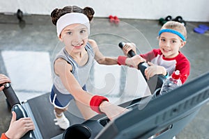 Adorable little girls in sportswear exercising on treadmill and smiling at camera in gym