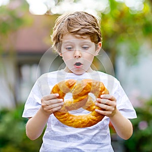 Adorable little kid boy eating huge big bavarian german pretzel. Happy blond child enjoying tasteful tratditional bread