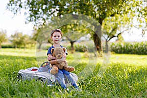 Adorable little kid girl sitting on big vintage old toy car and having fun with playing with big plush toy bear,