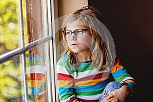 Adorable little preschool girl with eyeglasses sitting by the window. Thoughtful child looking out. Lonely kid.