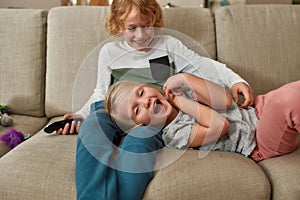 Adorable little siblings, boy and girl having fun, playing together while watching TV, cuddling on a sofa at home