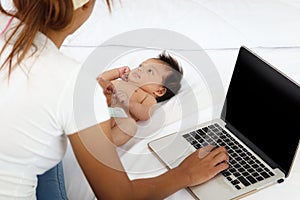 Adorable little sweet newborn baby girl lying on white bed near her mom using notebook laptop computer, cute infant with her busy