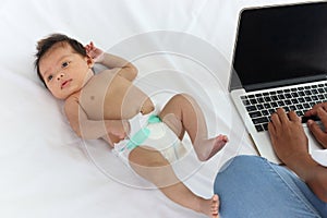 Adorable little sweet newborn baby girl lying on white bed near her mom using notebook laptop computer, cute infant with her busy