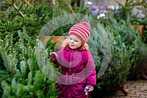 Adorable little toddler girl holding Christmas tree on market. Happy healthy baby child in winter fashion clothes