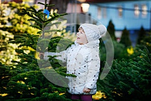 Adorable little toddler girl holding Christmas tree on market. Happy healthy baby child in winter fashion clothes