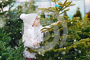 Adorable little toddler girl holding Christmas tree on market. Happy healthy baby child in winter fashion clothes