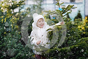 Adorable little toddler girl holding Christmas tree on market. Happy healthy baby child in winter fashion clothes