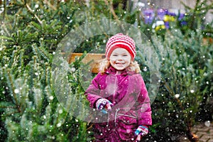 Adorable little toddler girl holding Christmas tree on market. Happy healthy baby child in winter fashion clothes