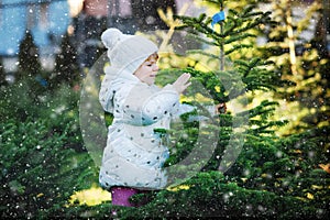 Adorable little toddler girl holding Christmas tree on market. Happy healthy baby child in winter fashion clothes choosing and