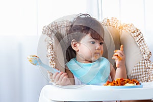 Adorable little toddler girl or infant baby eating delicious spaghetti food with tomato sauce on baby chair. Cute infant girl