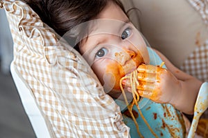Adorable little toddler girl or infant baby eating delicious spaghetti food with tomato sauce on baby chair. Funny cute infant