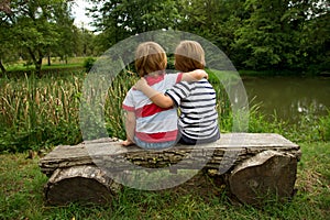 Adorable Little Twin Brothers Sitting on a Wooden Bench, Embracing Each Other and Looking at Beautiful Lake