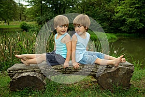 Adorable Little Twin Brothers Sitting on a Wooden Bench, Smiling and Looking at Each Other Near the Beautiful Lake