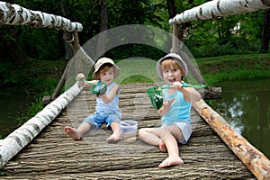 Adorable Little Twin Brothers Sitting on a Wooden Bridge and Holding a Fishnet Full of Fish at the Lake