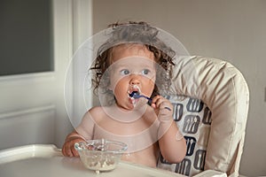 Adorable long haired toddler sitting in a baby high chair and eating porridge indoor.