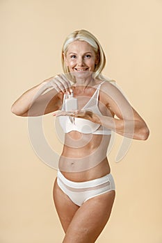 Adorable mature woman in underwear holding and opening white jar of skin care cream or lotion, standing  over