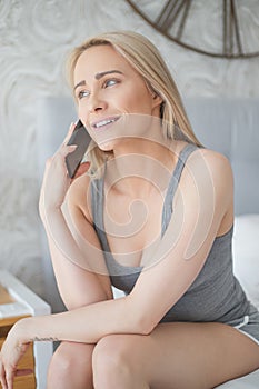 Adorable middle aged blonde woman sitting on bed and talking on mobile phone