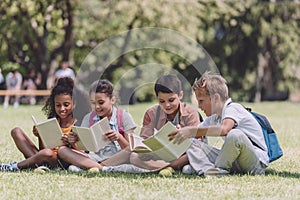 Adorable multicultural schoolkids sitting on lawn and reading books