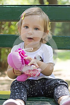 Adorable one year old baby girl sitting on a bench with a doll i
