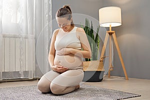 Adorable pleased pregnant woman wearing beige clothing sitting on floor in living room touching and looking at her tummy restring photo