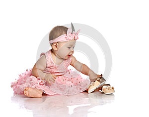 Adorable little baby girl in stylish pink princess dress, decorative crown and barefoot sitting on floor and playing with shoes