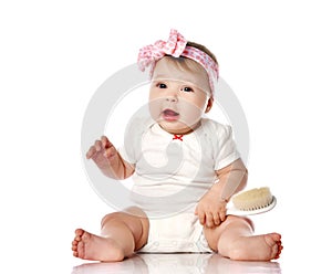 Adorable positive little baby girl in white body, hair bow decoration sitting on floor, smiling and holding brush in hand