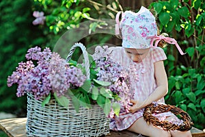 Adorable preschooler girl in pink plaid dress making lilac wreath in spring sunny garden