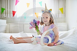 Adorable preschooler girl in red dress sitting on bed with pink hyacinths and white wooden number six. Sixth birthday concept. Kid