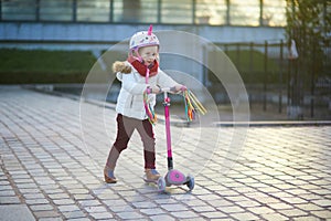 Adorable preschooler girl riding her scooter in a city park on sunny spring day