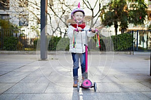 Adorable preschooler girl riding her scooter in a city park on sunny spring day