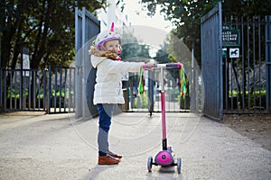 Adorable preschooler girl riding her scooter in a city park on sunny spring day