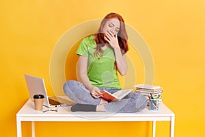 Adorable red haired tired sleepy woman sitting on her desk with books and computer, keeps eyes closed, covering her mouth while