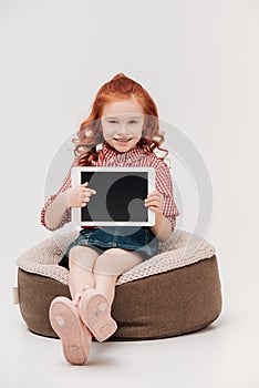 adorable redhead child smiling at camera and pointing at digital tablet with blank screen