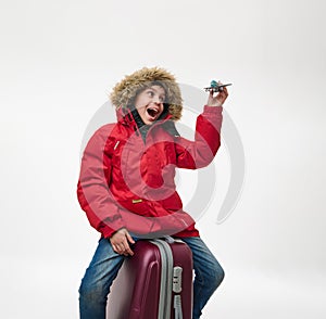Adorable school boy in warm red down jacket with hood, sitting on a suitcase and playing with a toy model of an airplane,
