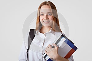 Adorable school girl with blue eyes, shining smile, wears elegant white shirt, carries books and rucksack, has long hair, prepares