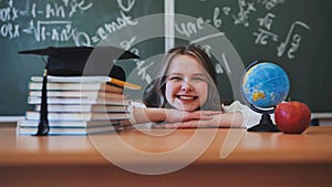 Adorable school girl posing at her desk against a background of blackboard, books, globe and graduation cap.