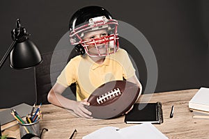 adorable schoolboy in american football helmet sitting at table with ball books lamp colour pencils textbook on grey photo