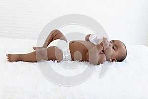 Adorable a sixmonth crawling African American baby lying on fluffy white rug, happy sweet little girl kid holding milk bottle and