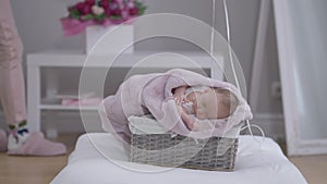 Adorable sleeping infant in pink blanket in baby basket with woman putting flowers at background on table. Wide shot