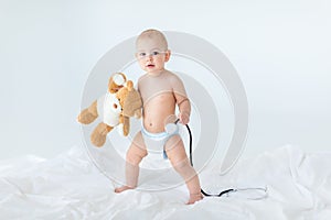 Adorable small baby boy standing on bed and holding stethoscope with teddy bear