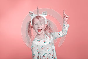 adorable smiling Caucasian blonde girl in white dress with unicorn headband horn on pink coral background