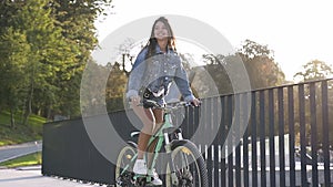 Adorable smiling hot girl in jeans clothes riding on bicycle near metal fence