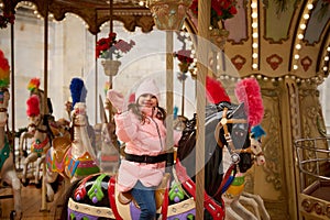 Adorable smiling kid girl waving hello while riding on merry go round carousel horse at Christmas winter market outdoor