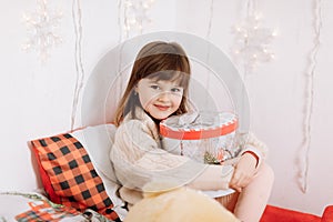 Adorable smiling little child girl is holding box with Xmas present and looking at camera in cozy bedroom, background of