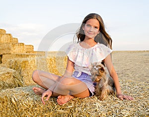 Adorable smiling little girl child sitting on a hay rolls in a wheat field with her small dog pet yorkshire terrier