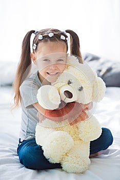 Adorable smiling littlegirl sitting on a bed hugging a white teddy bear